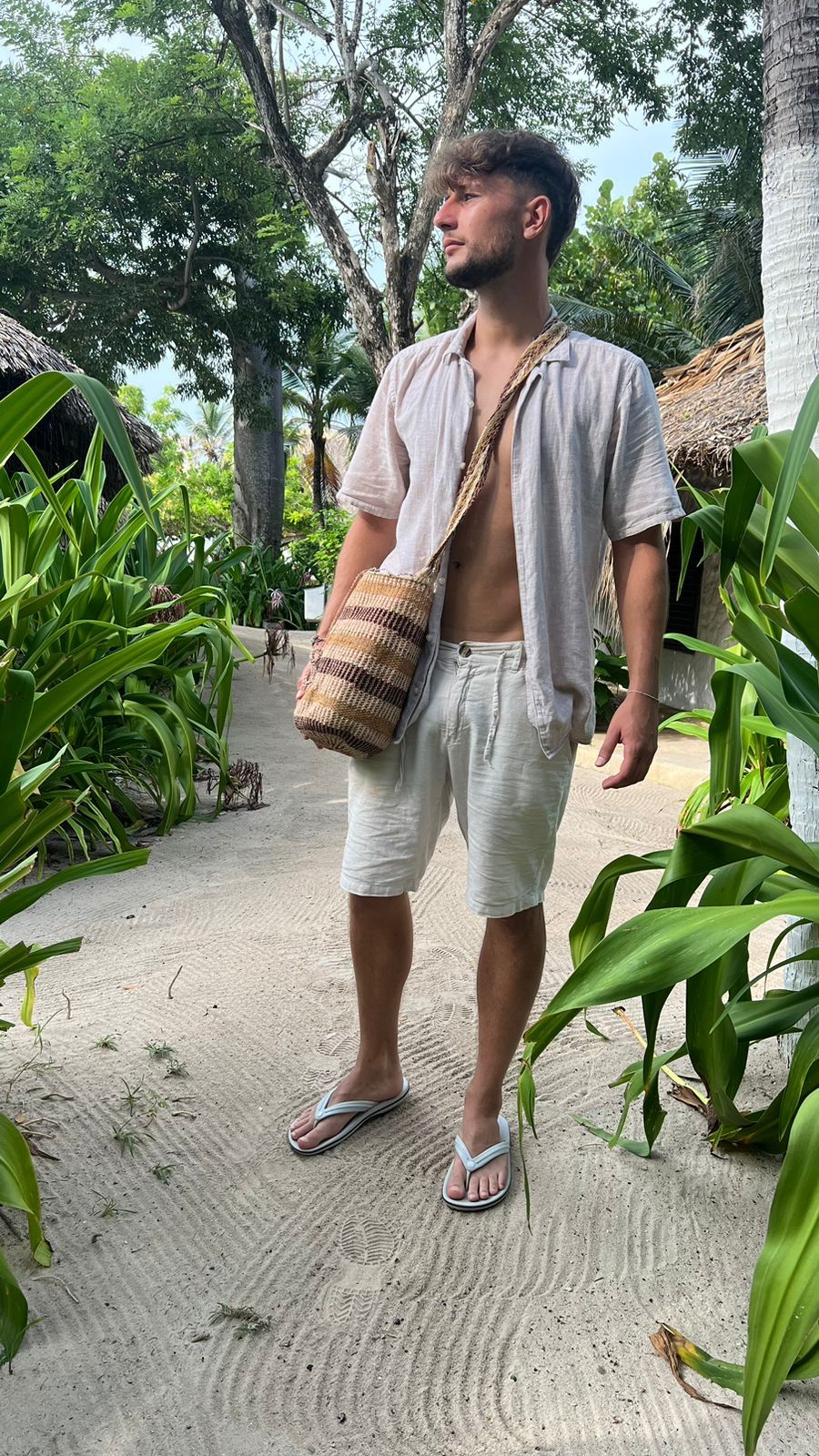 Man wearing a relaxed open shirt and white shorts, carrying a handmade Indigenous mochila bag in a tropical beachside setting, styled as a sustainable tote bag for men’s ethical fashion.