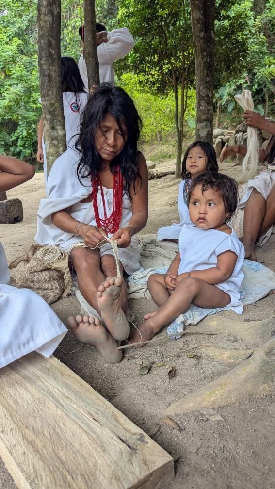 Indigenous Kogui elder Johanna hand-weaving a mochila bag while seated on the ground in Tayku, Colombia, with young children sitting near her, surrounded by trees and natural fiber.