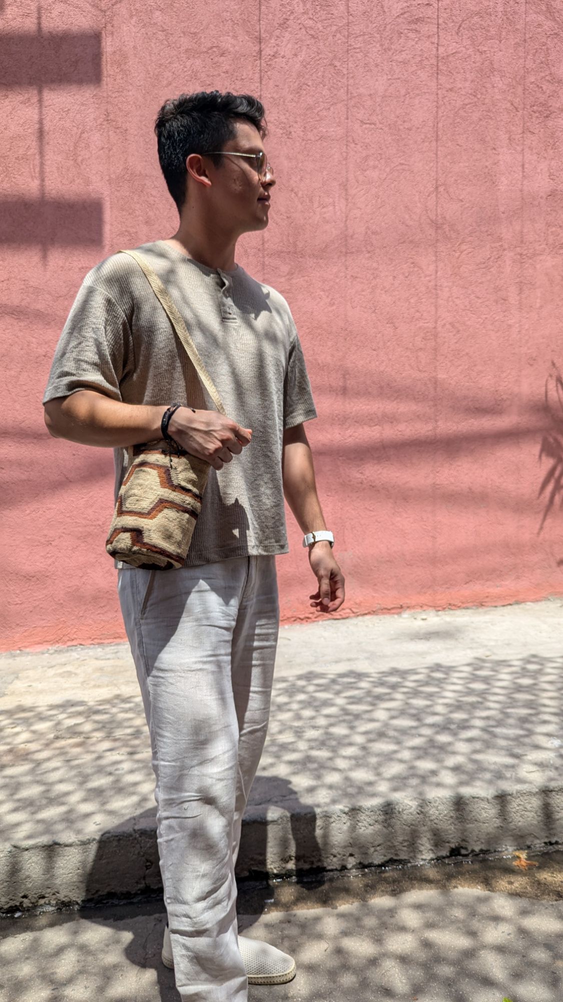 Man wearing a handwoven mochila bag with neutral-toned linen outfit, standing against a pink wall in sunlight, representing slow fashion and Indigenous craftsmanship.