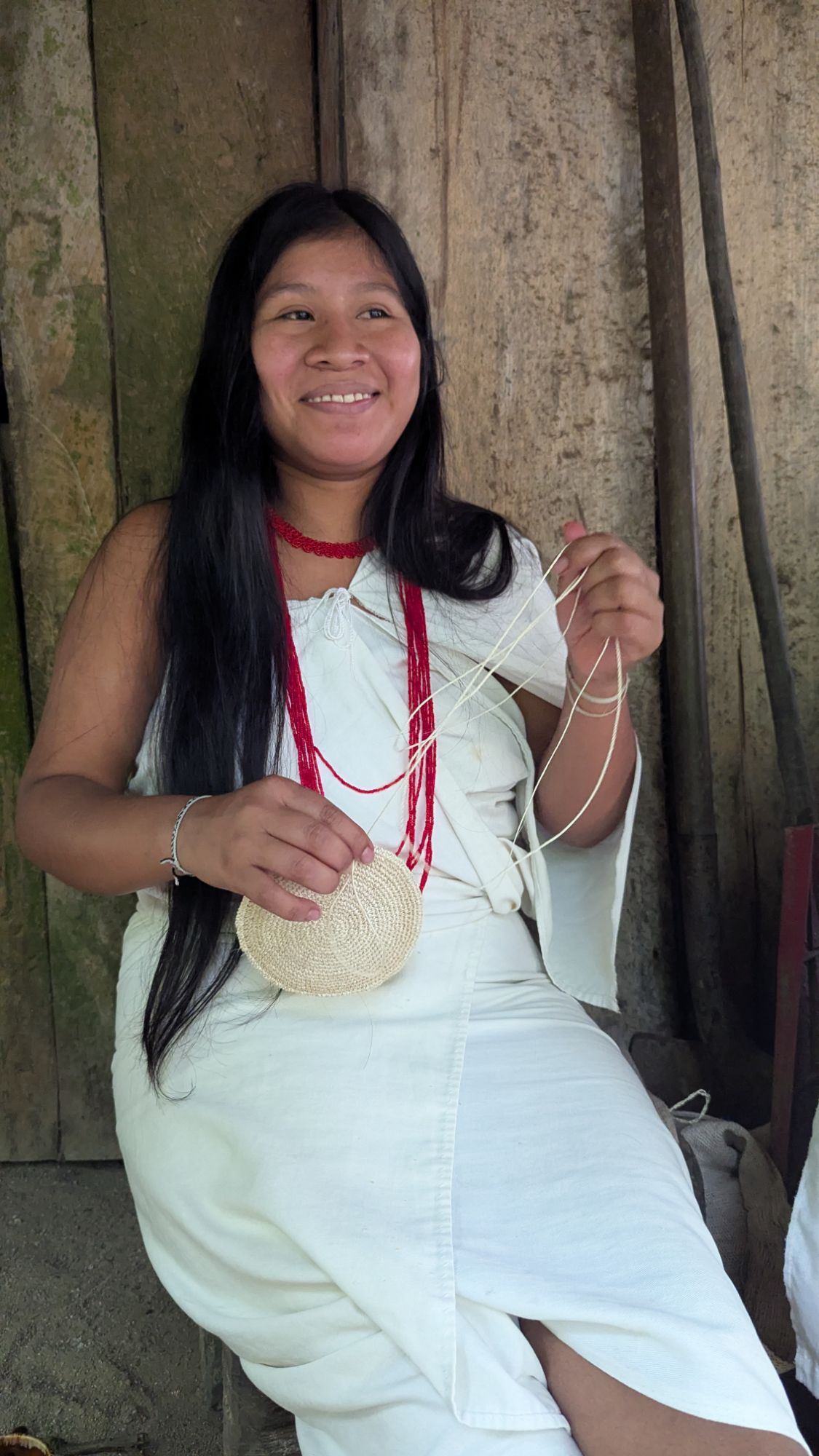 Portrait of Indigenous Kogui artisan Francisca seated outdoors, smiling as she hand-weaves a mochila bag using natural fiber, wearing traditional white clothing and red necklace in Tayku, Colombia.